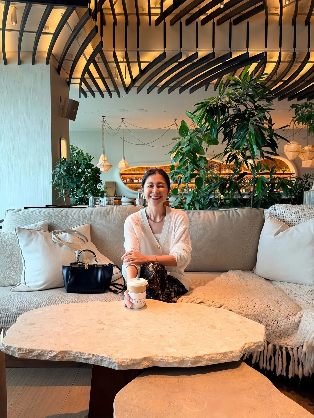 １Hotel Tokyo 38階、女性が石製のテーブルに座り、コーヒーカップを持っています。白いソファに黒いハンドバッグが置かれ、背景には緑の植物とモダンな照明が配置されています。