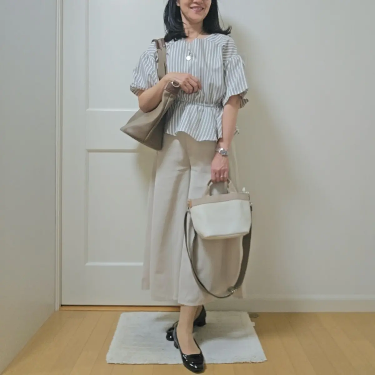 白い壁に立っている女性で、グレーのストライプのプルオーバーとベージュのワイドパンツを着用。左肩に大きめのベージュのトートバッグをかけ、右手には小さめの白いショルダーバッグを持っています。