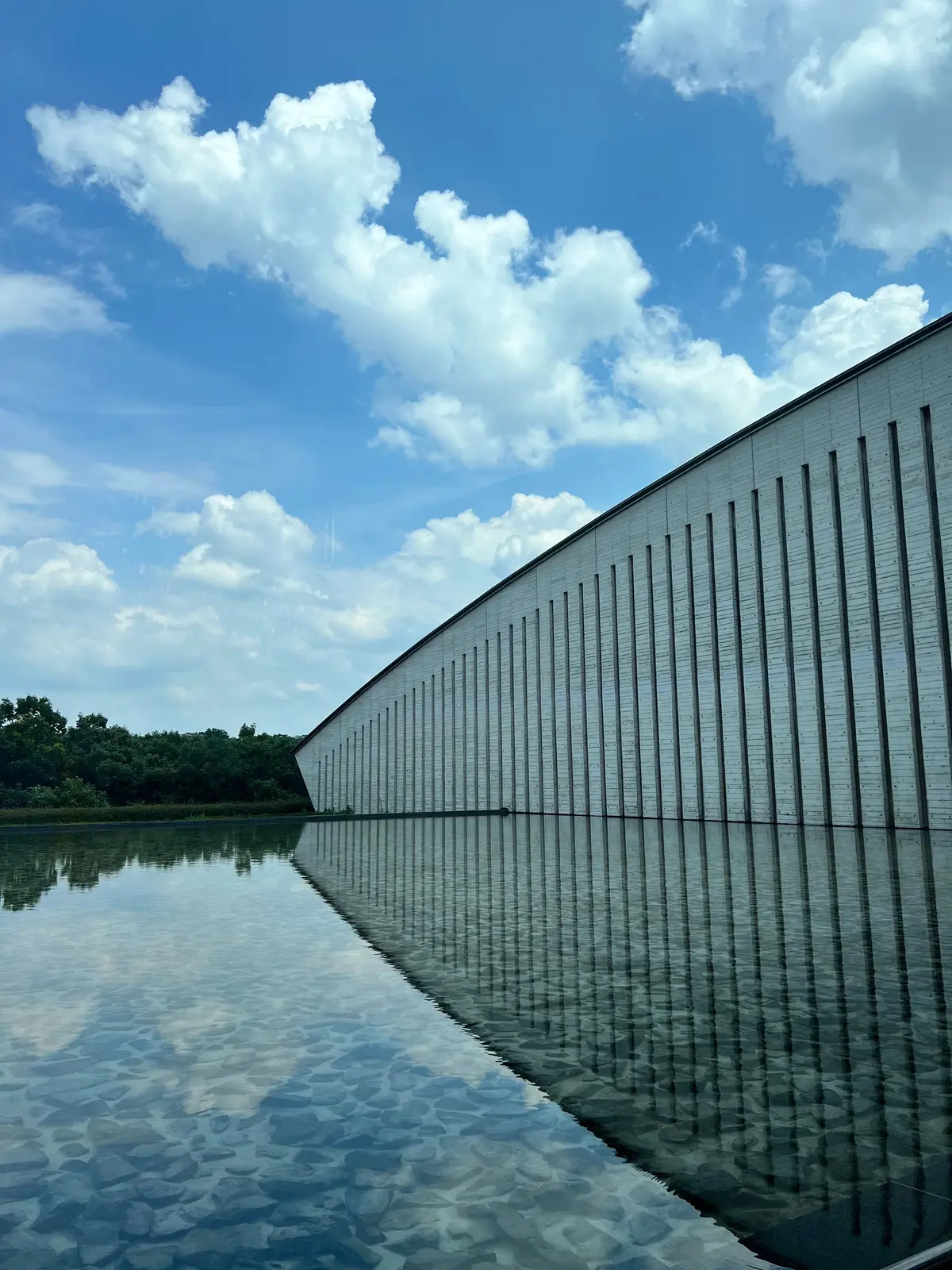 奥田元宋・小由女美術館。水面に建物が映る風景。グレーの壁面は縦の溝が刻まれ、空と雲が青く広がる。左右対称の構図で、水面には空と建物の反射が美しい。