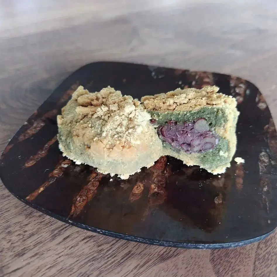 断面されたお菓子。緑色の生地で紫色の餡が包まれ、表面には茶色い粉がふられている。木製の皿に置かれている。