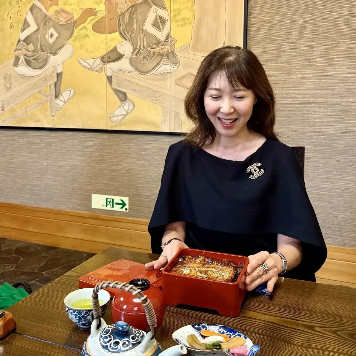 笑顔の女性が、漆塗りの箱に入った魚料理を手に持っている。背景には日本らしい絵画が飾られ、テーブルにはお茶や小皿が並んでいる。