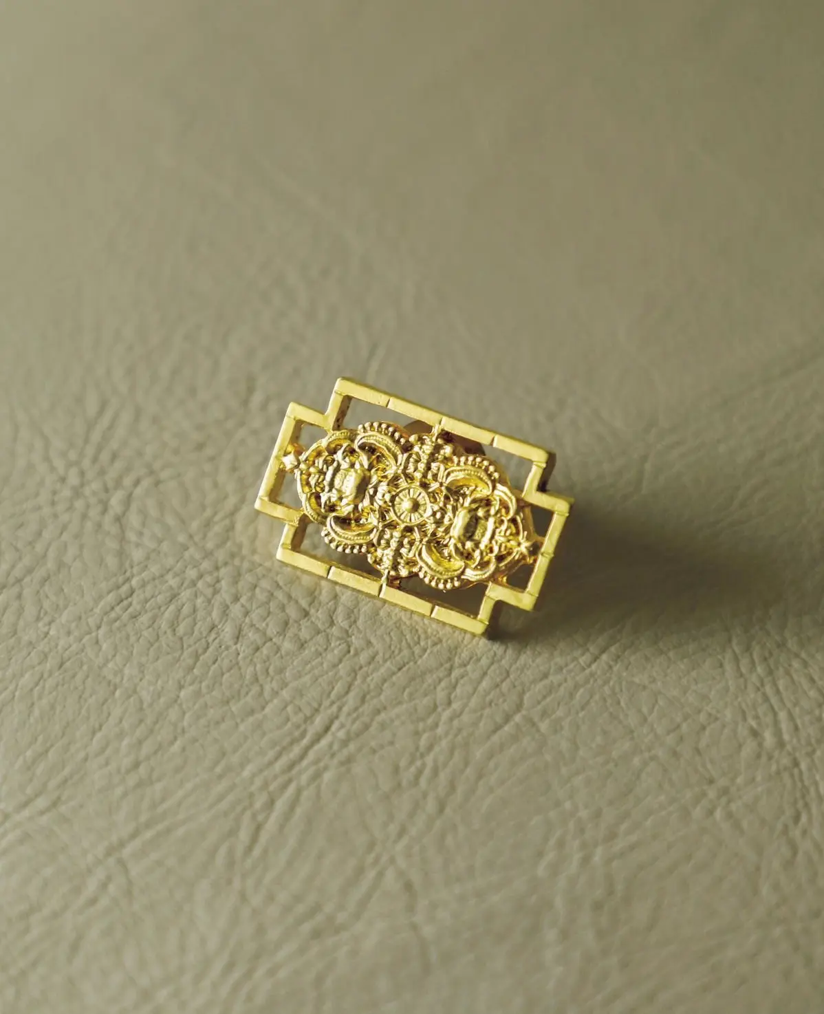 金色の装飾的なブローチ。長方形の枠の中に、繊細な模様が施されたデザインが配置されている。表面は光沢があり、背景の質感のあるベージュの布の上に置かれている。