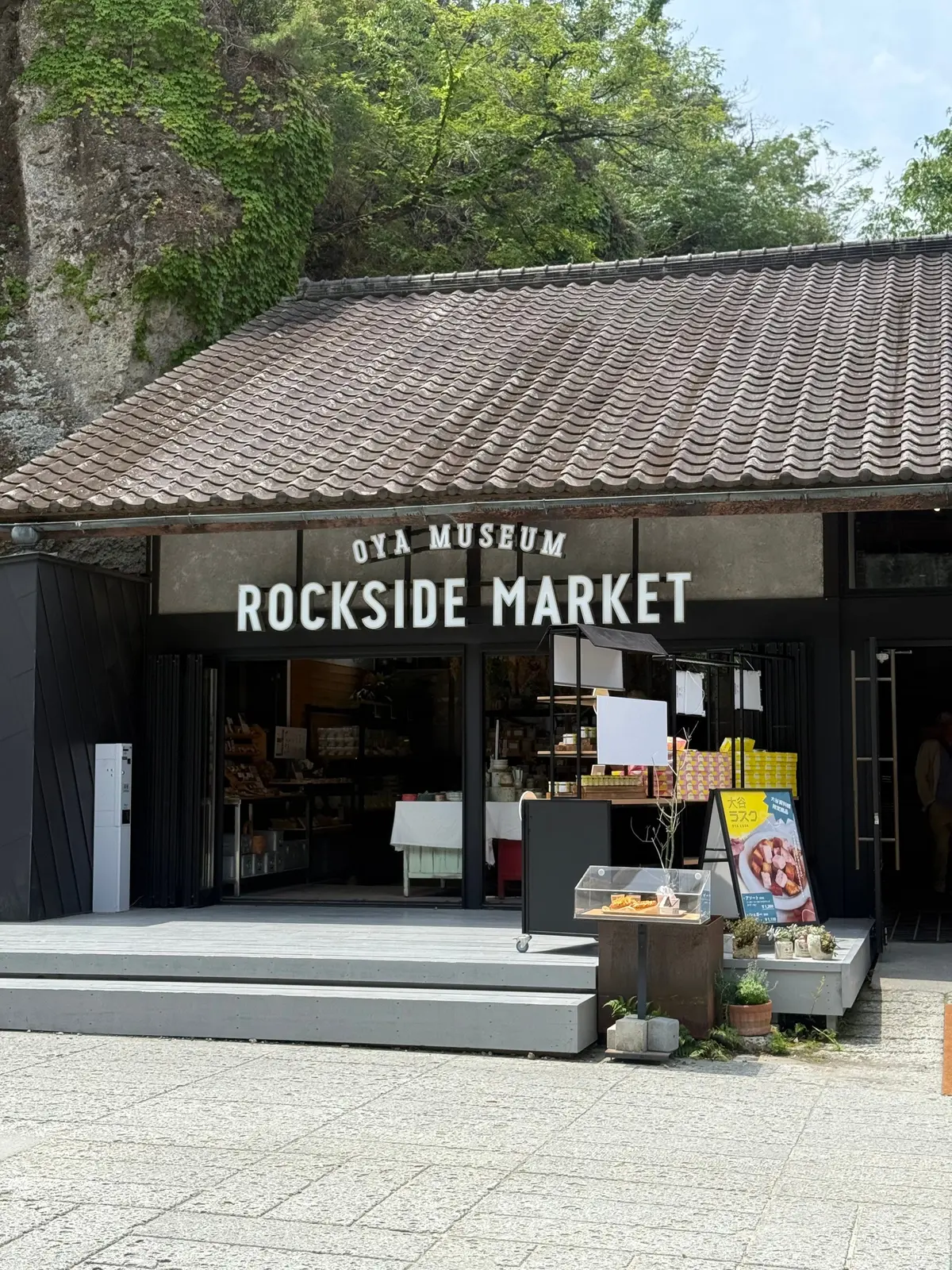 OYA MUSEUMの ROCKSIDE MARKETエントランス。瓦屋根の建物にガラスのドアがあり、店内には商品が並ぶ。外にはテーブルと椅子、花鉢が置かれている。