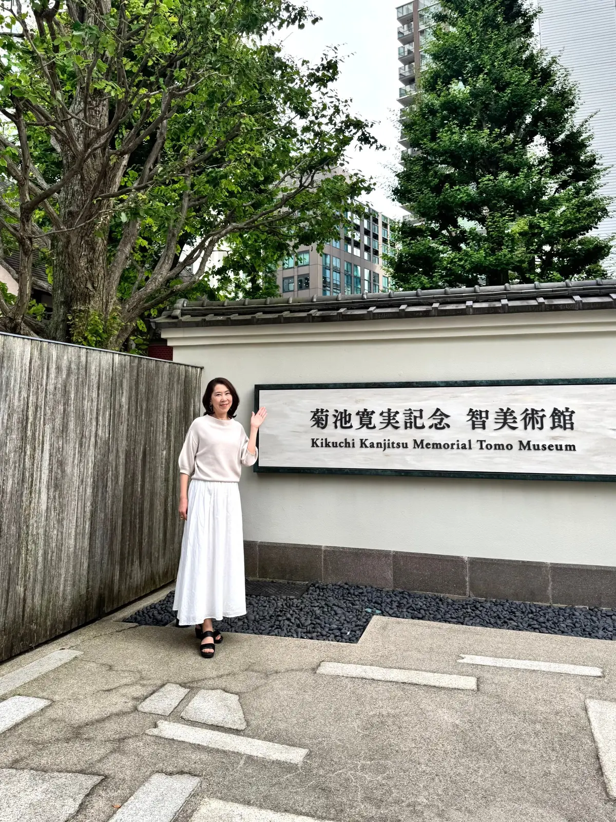 白いトップスとスカートを着た女性が、菊池寛実記念智美美術館の前で手を振っている。背景には緑豊かな木々と近代的な建物が見える。