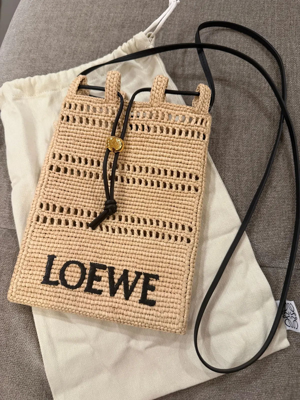 ベージュの編みかごバッグで「LOEWE」の文字入り、黒のストラップと金色のチャームが付いたデザイン