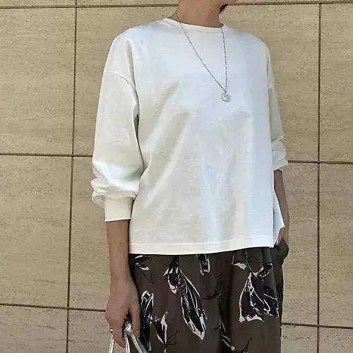 白いゆったりとした長袖カットソーを着た女性。茶色のタイル壁を背景に、柄入りのボトムスを着用。シンプルなシルバーネックレスを身につけている。