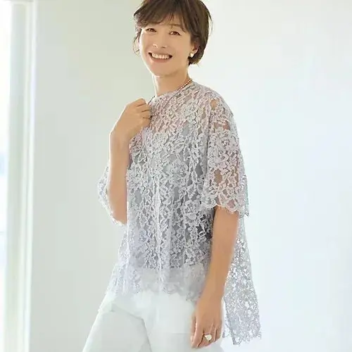上品なレース素材のグレーのトップスを着用した女性。白いパンツを合わせ、微笑みながらこちらを見ている。背景は白い壁で、窓からの光が差し込んでいる。