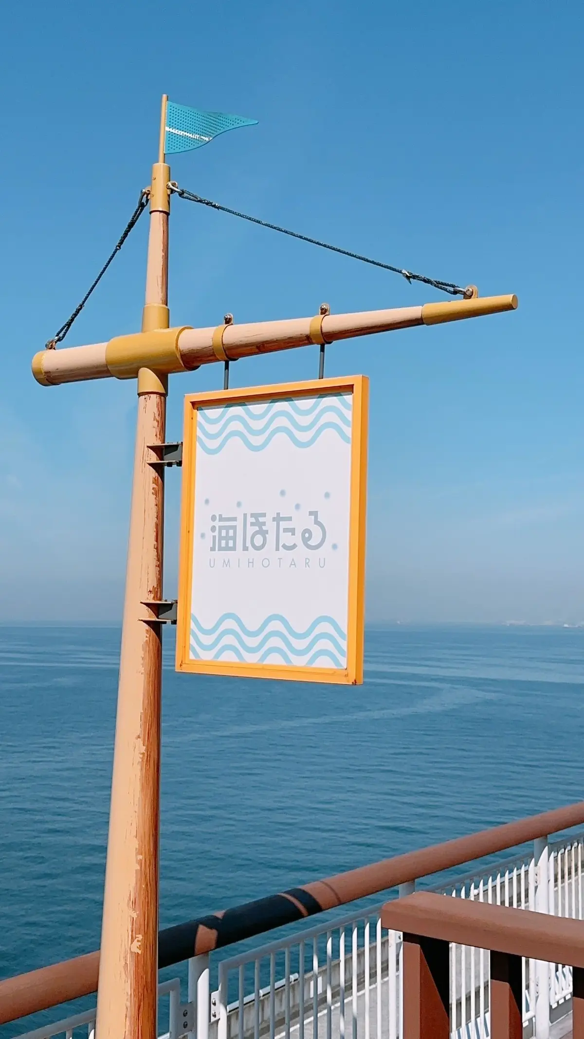 青い空と海を背景に、オレンジ色の柱に取り付けられた「UMI HOTARU」と書かれた看板。波のイラストと小さな旗が飾られている。
