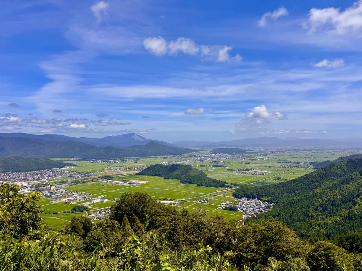 青空を背景に、緑豊かな田園風景と山々が広がる眺望。手前には木々が生い茂り、遠景には集落と連なる山々が連なる。