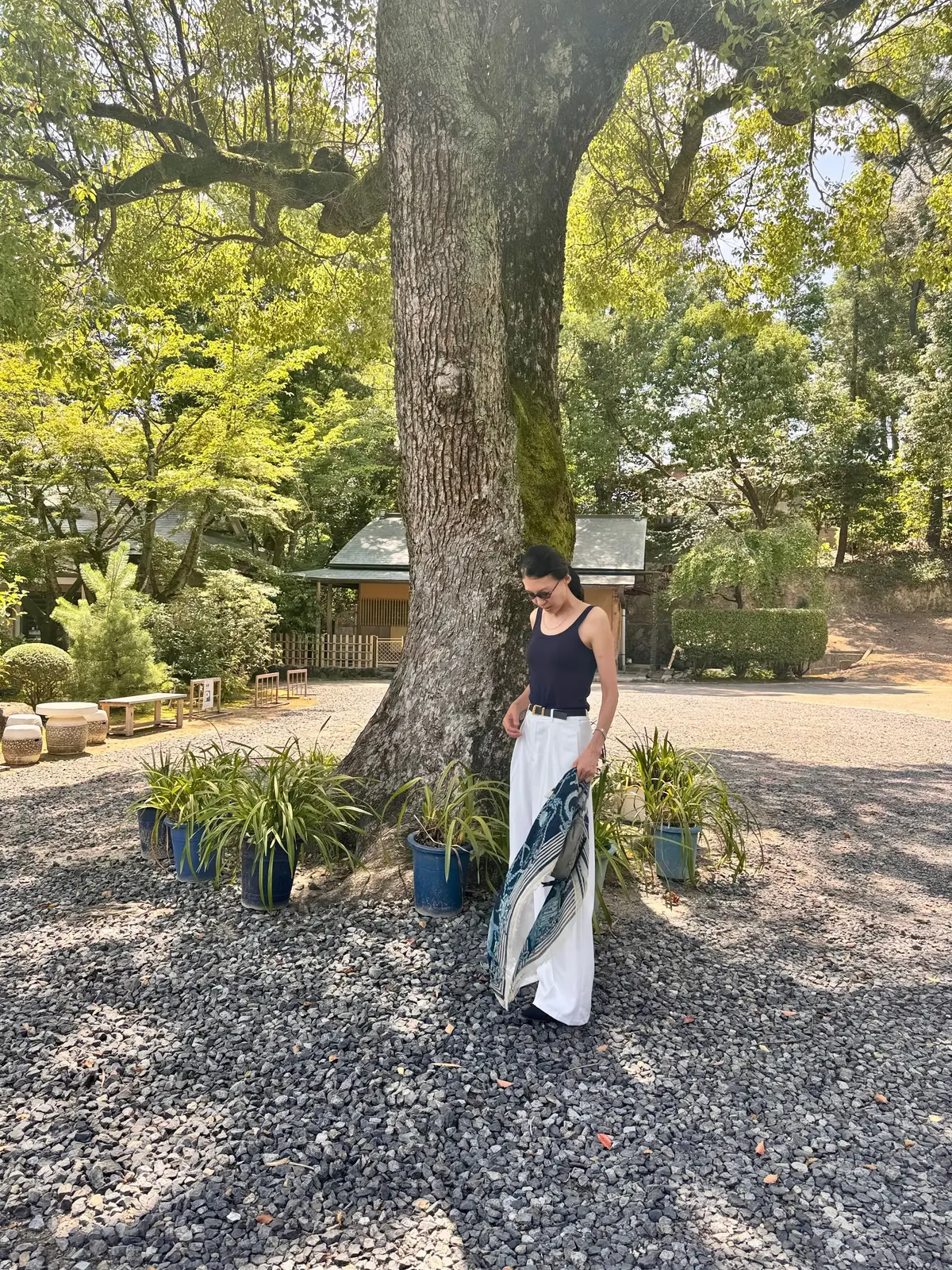 白いパンツと黒いタンクトップを着た女性が、大きな木のそばに立っている。足元には青い植木鉢に入った植物が並び、背景には木々が生い茂る庭園が見える。
