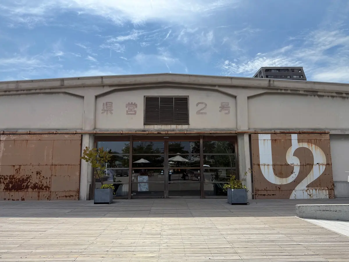 白っぽいコンクリートの建物正面。中央に「県営2号」の文字、左右に錆びた鉄板の装飾がある。ガラス窓の向こうに緑が見える。手前に木製のデッキがある。尾道、ONOMICHI U2。デニムの街で尾道観光、広島。