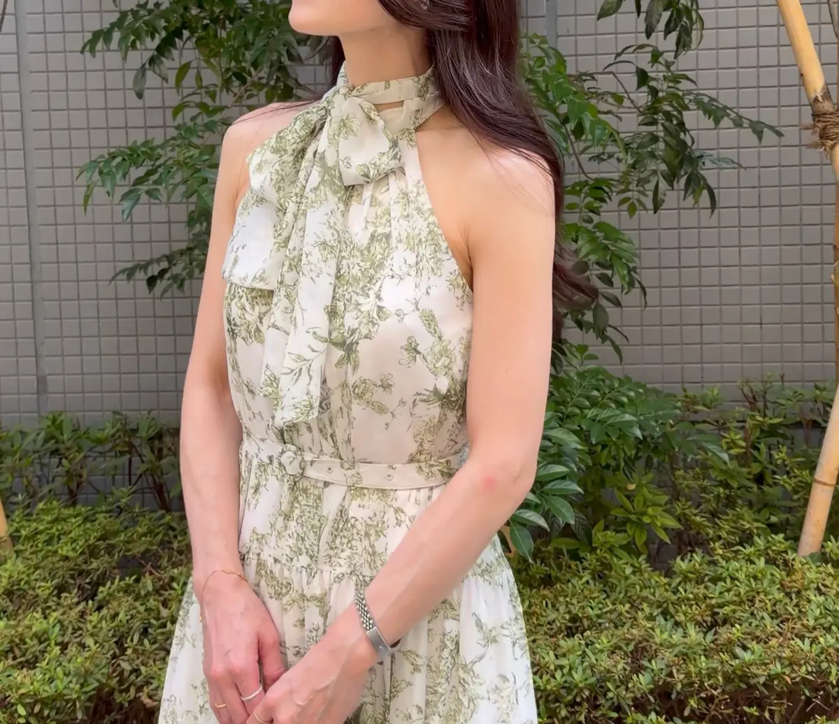 クリーム色地に緑色の花柄がプリントされた、ふんわりとしたロングワンピースを着た女性。首元にリボン結びのディテールがあり、右手にパステルグリーンの小さなバッグを持つ。背景には緑の植栽とグレーの壁がある。