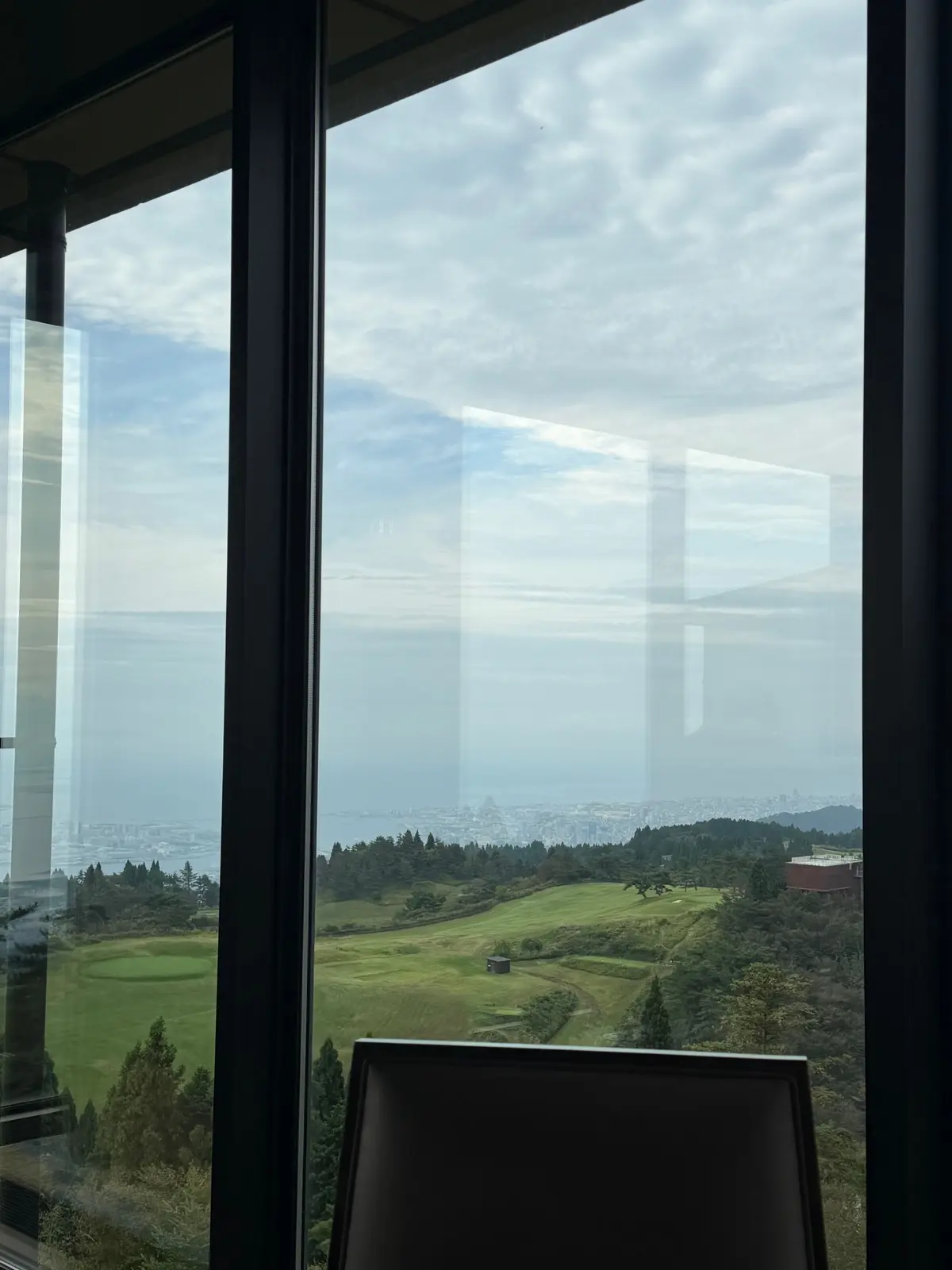 高台から見下ろす、緑豊かな丘陵地と遠くの都市の風景。窓枠が前景にあり、雲が広がる空が背景に見える。