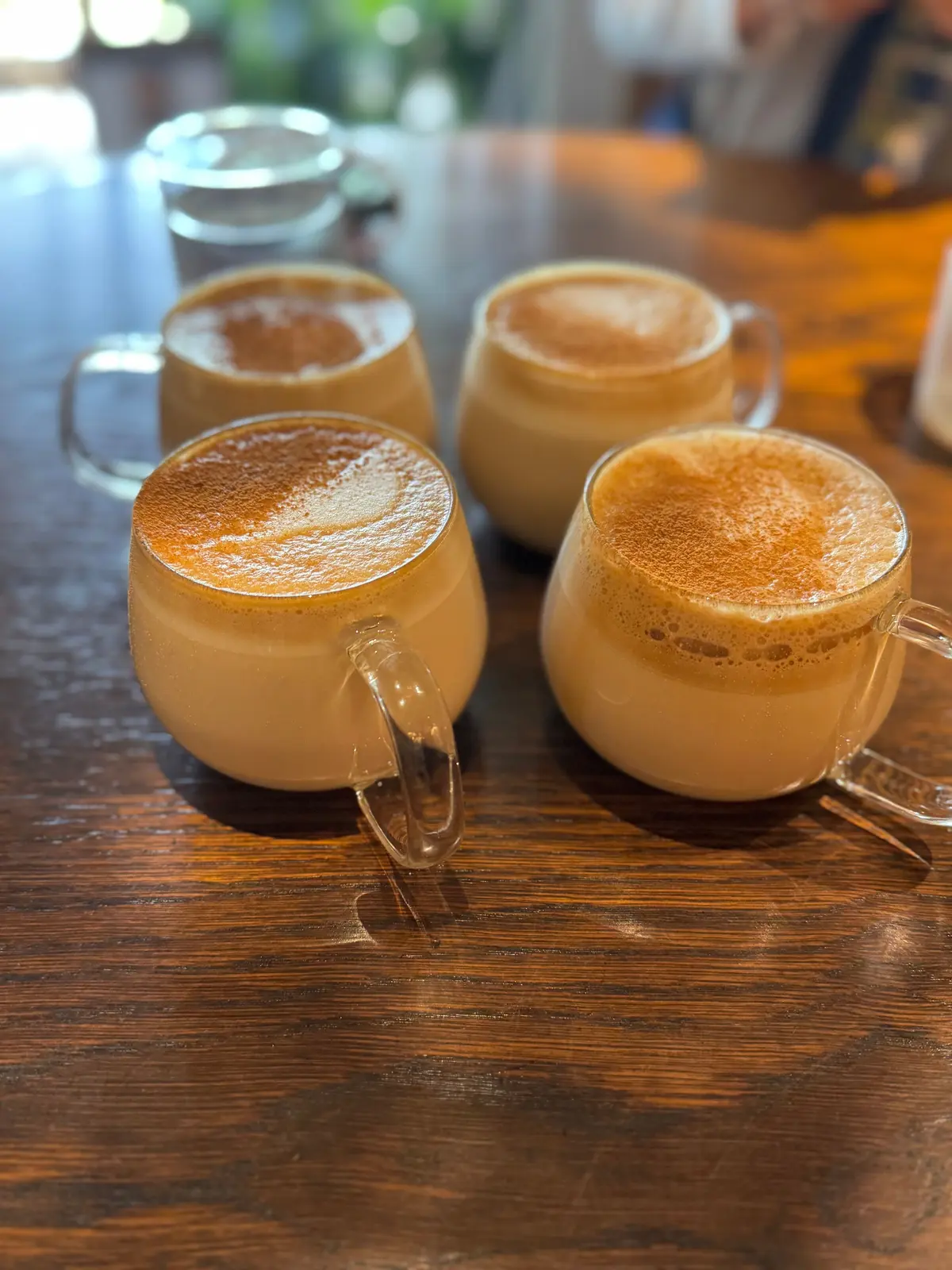 木製テーブルに置かれた4つのガラスカップ、温かい飲み物とふわふわのフォームが特徴