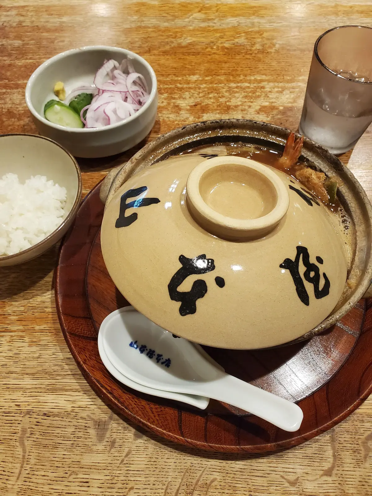 木製のトレイに、陶器の鍋に蓋をした一品料理、米、野菜の小鉢、グラスが並んでいる