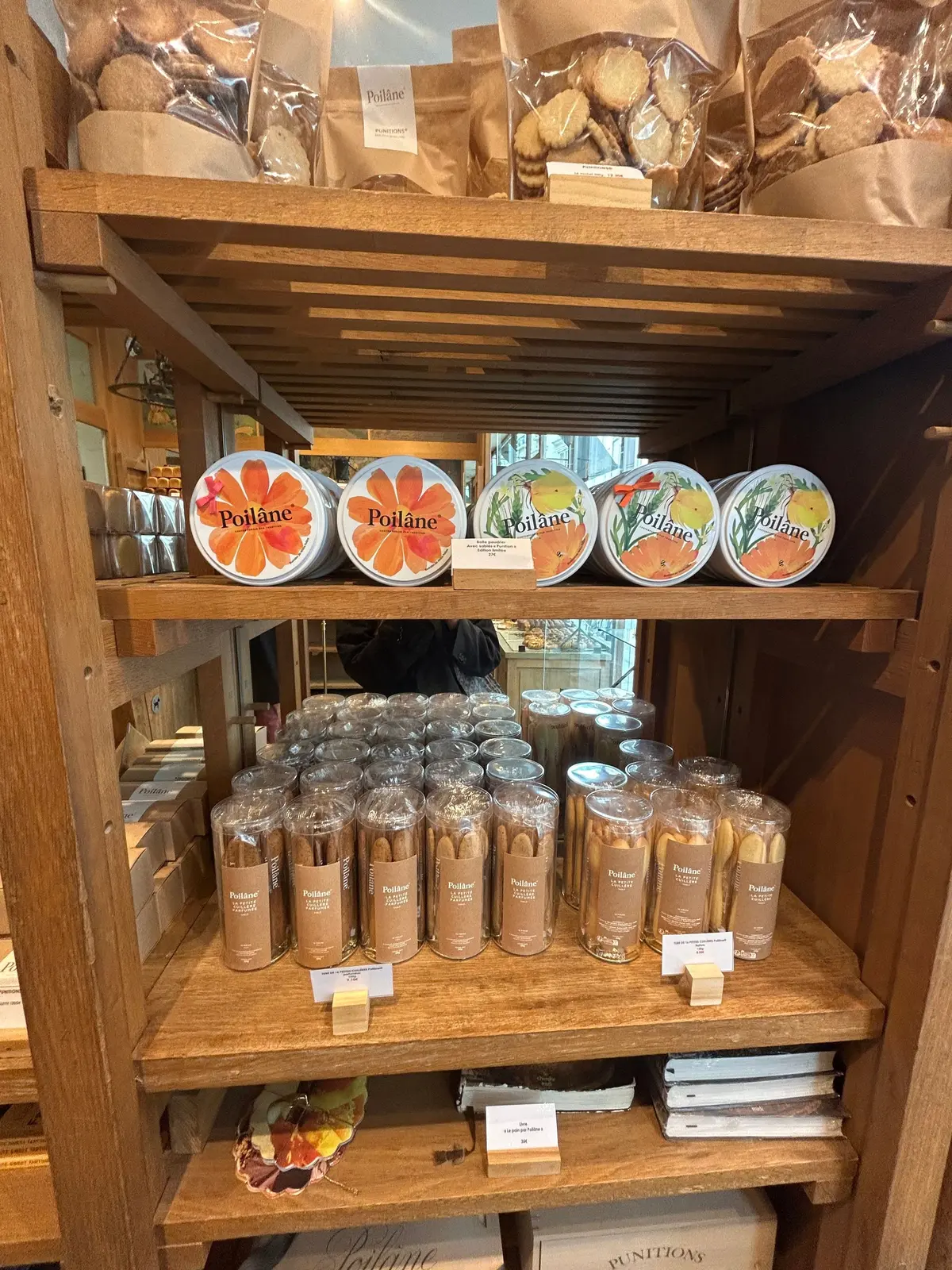 木製の棚に「Poilâne」の焼き菓子と缶詰が並ぶ。棚の上段には紙袋に入ったパンの塊と缶詰が置かれ、中段には花模様の缶と透明なプラスチックカップに入った焼き菓子が並ぶ。下段には本の山と小型の箱が置かれている。