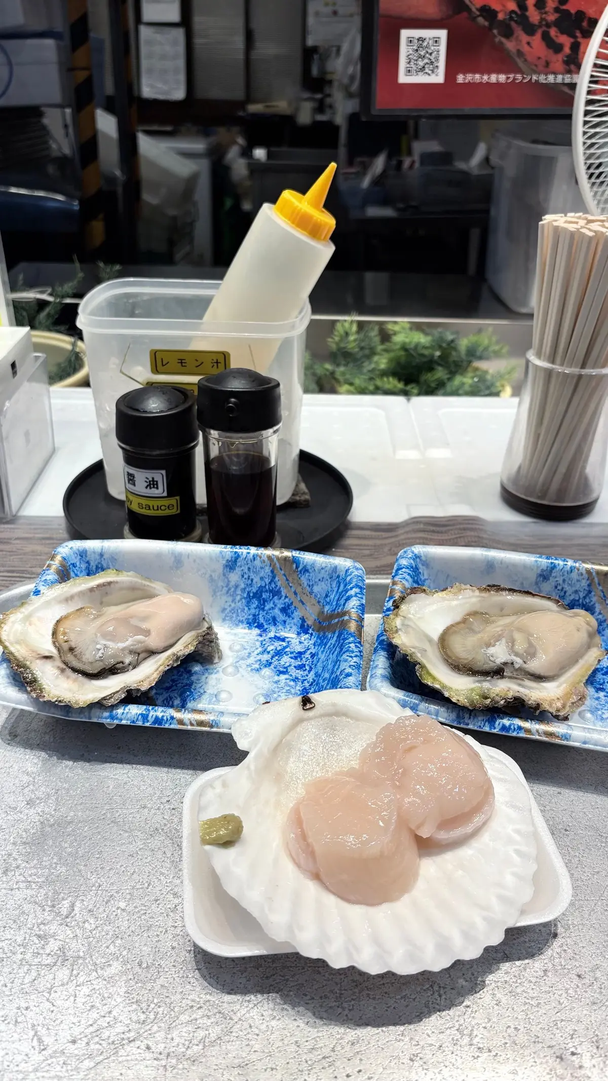 青い紙の上に盛り付けられたカキと、薄切りになった魚介類。レモン汁、醤油、割り箸が近くに置かれている。背景には、ステンレス製のカウンターと調理器具が見える。