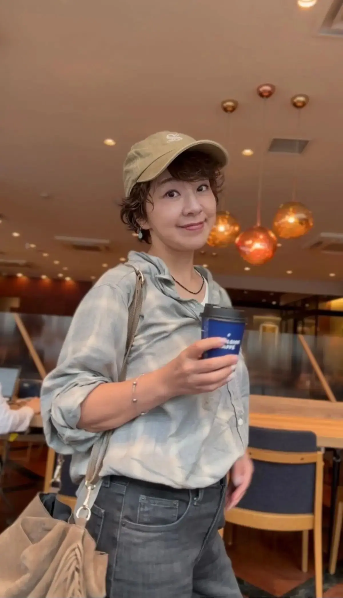 カジュアルな服装の若い男性が、カフェでコーヒーを持ちながら微笑んでいる。キャップを被り、グレーのシャツとデニムを着用。背景には、おしゃれな照明器具が並ぶカフェの様子。