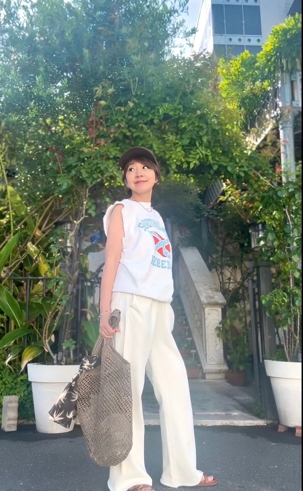 女性が白いワイドパンツと白いTシャツ、キャップをかぶり、屋外で立っている。左手に編み目のトートバッグを持っている。背景には緑の植物と白い花壇がある。
