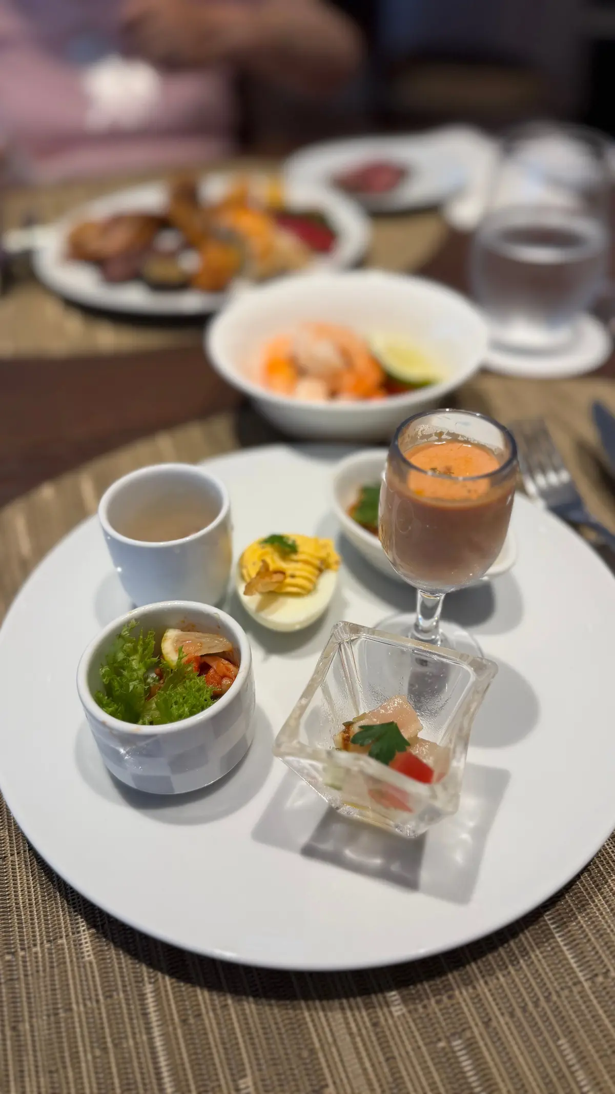 白い皿に盛り付けられた、様々な小皿料理。エビと野菜のサラダ、スープ、パスタ、ゼリー、そして白い器に入った料理が並んでいる。背景には他の料理と飲み物が写っている。