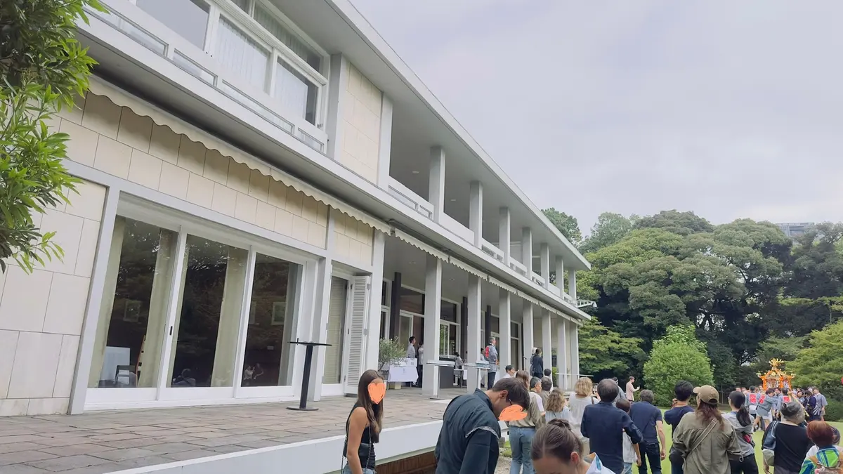 白とベージュの近代的な建物と、その前庭に集まる人々。大きな窓と柱が特徴的で、テラス席がある。背景には緑豊かな木々が見える。