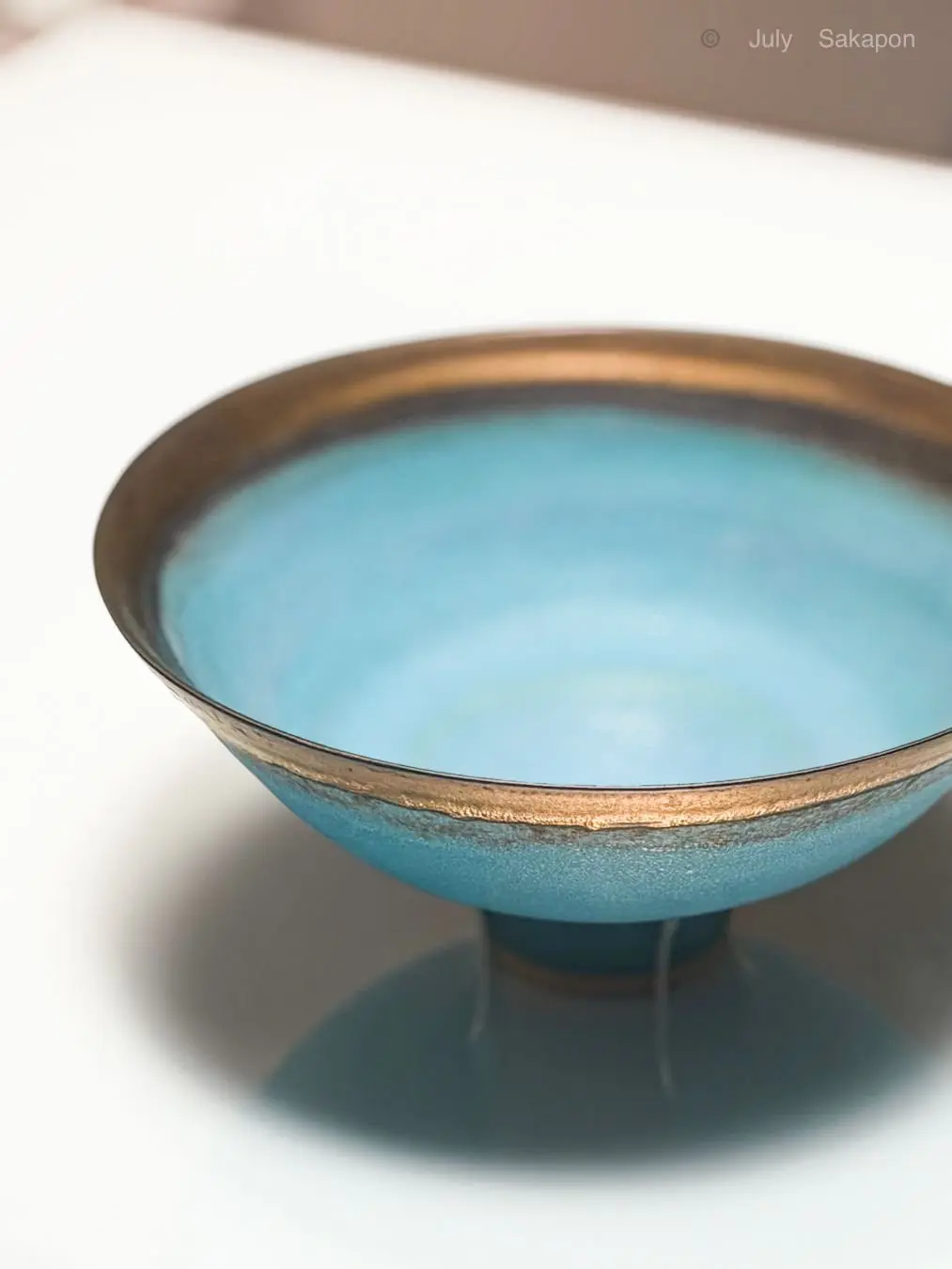 青色の釉薬を施した浅い器。金色の縁取りが施され、金属製の脚で支えられている。白いテーブルの上に置かれ、影を落としている。