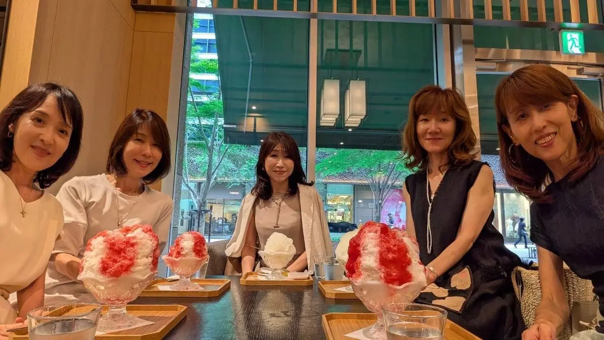 5人の女性がテーブルを囲み、かき氷を前に笑顔を見せている。かき氷はピンクと白の層でできており、ガラスの器に入っている。背景には窓から緑の木々が見える。