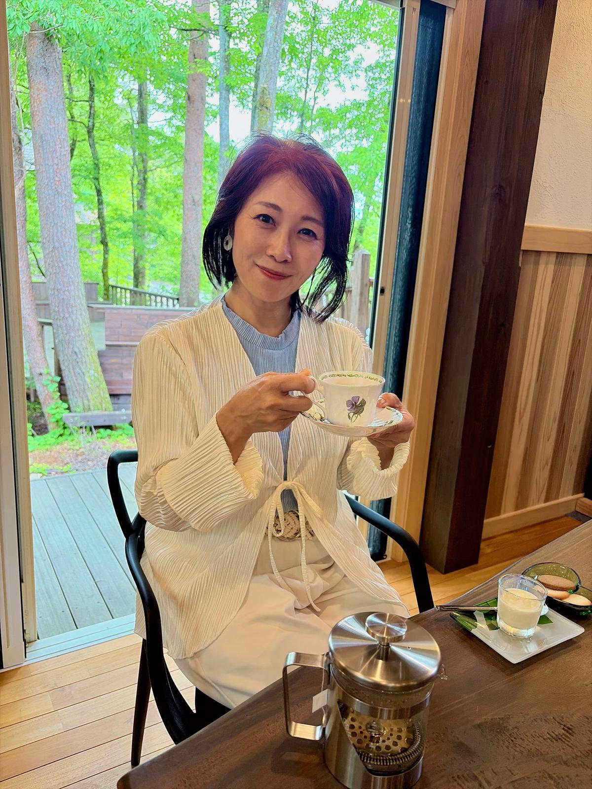 女性が窓辺のテーブルでティーカップに茶を注いでいる。木製のテーブルにはミルクとクッキーも並んでいる。背景には緑豊かな木々が広がる。