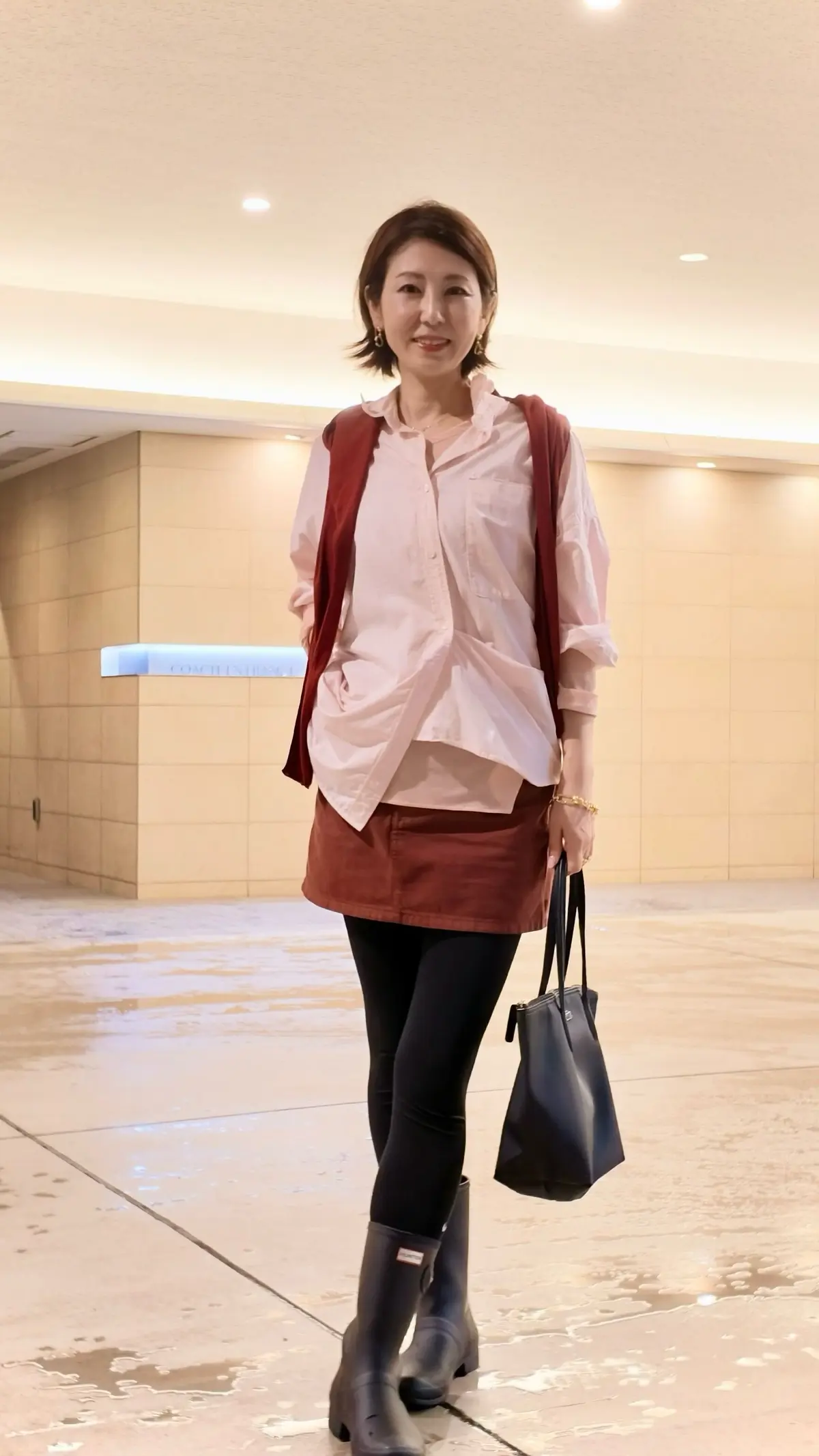 ブラウンのブーツと黒のタイツを履いた女性が、赤いミニスカートと白いシャツ、赤いカーディガンを着用して立っている。背景には「COACH ENTRANCE」と書かれた案内板が見える。左手にブルーのハンドバッグを持っている。