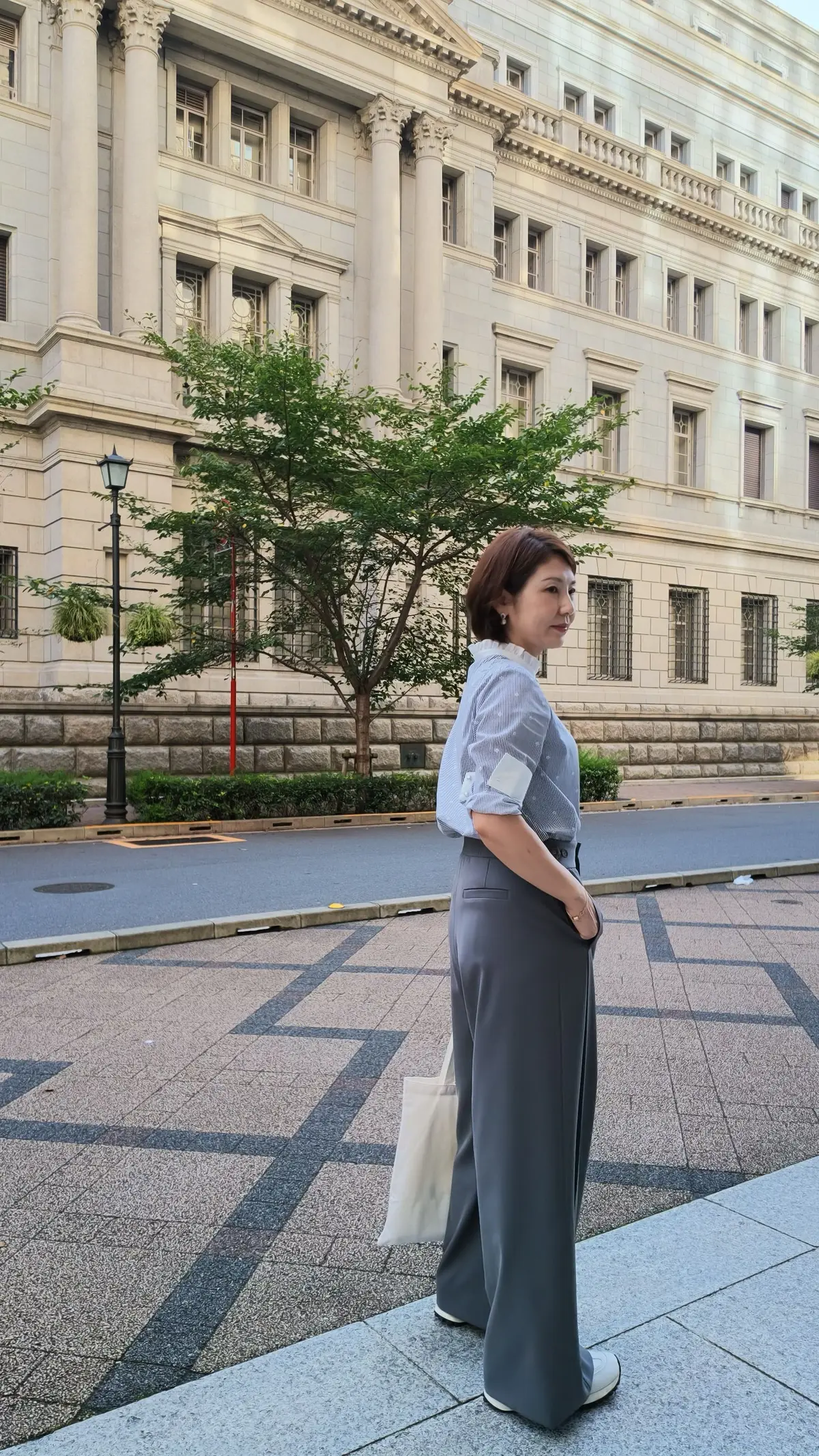 グレーのパンツとシャツを着た女性が、クラシカルな建物と緑の木を背景に立っている。右手に白いバッグを持っている。