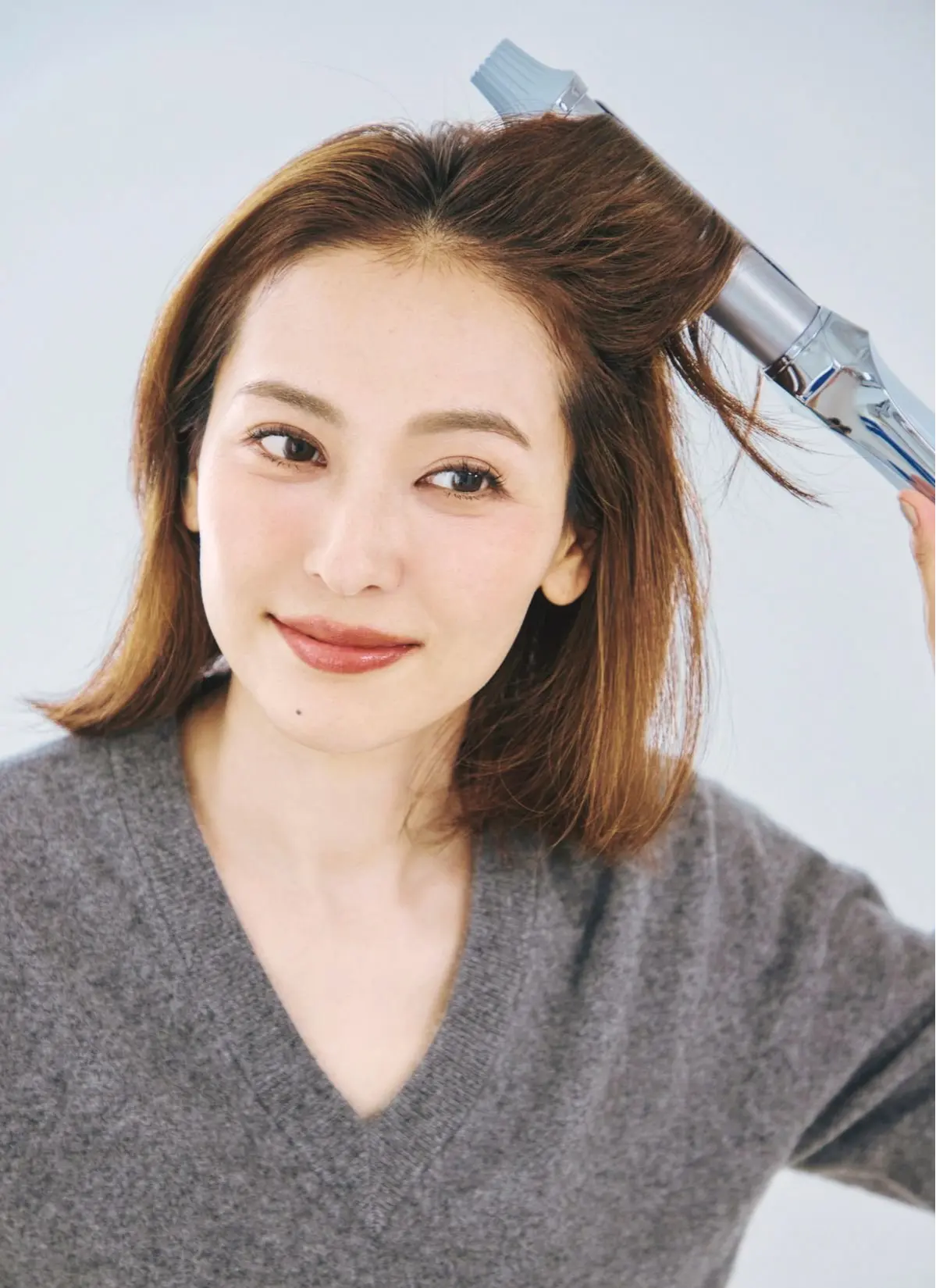 茶髪の女性が、シルバーのヘアードライヤーで前髪を乾かしている。グレーのセーターを着て、白い背景の前で微笑んでいる。