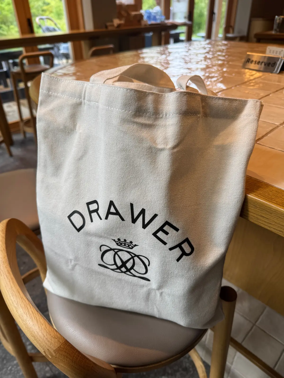 白地のトートバッグが椅子に置かれている。バッグ正面中央に黒色で「DRAWER」の文字と、その下に王冠をモチーフにしたロゴマークが刺繍されている。背景には木製のテーブルと椅子が見える。