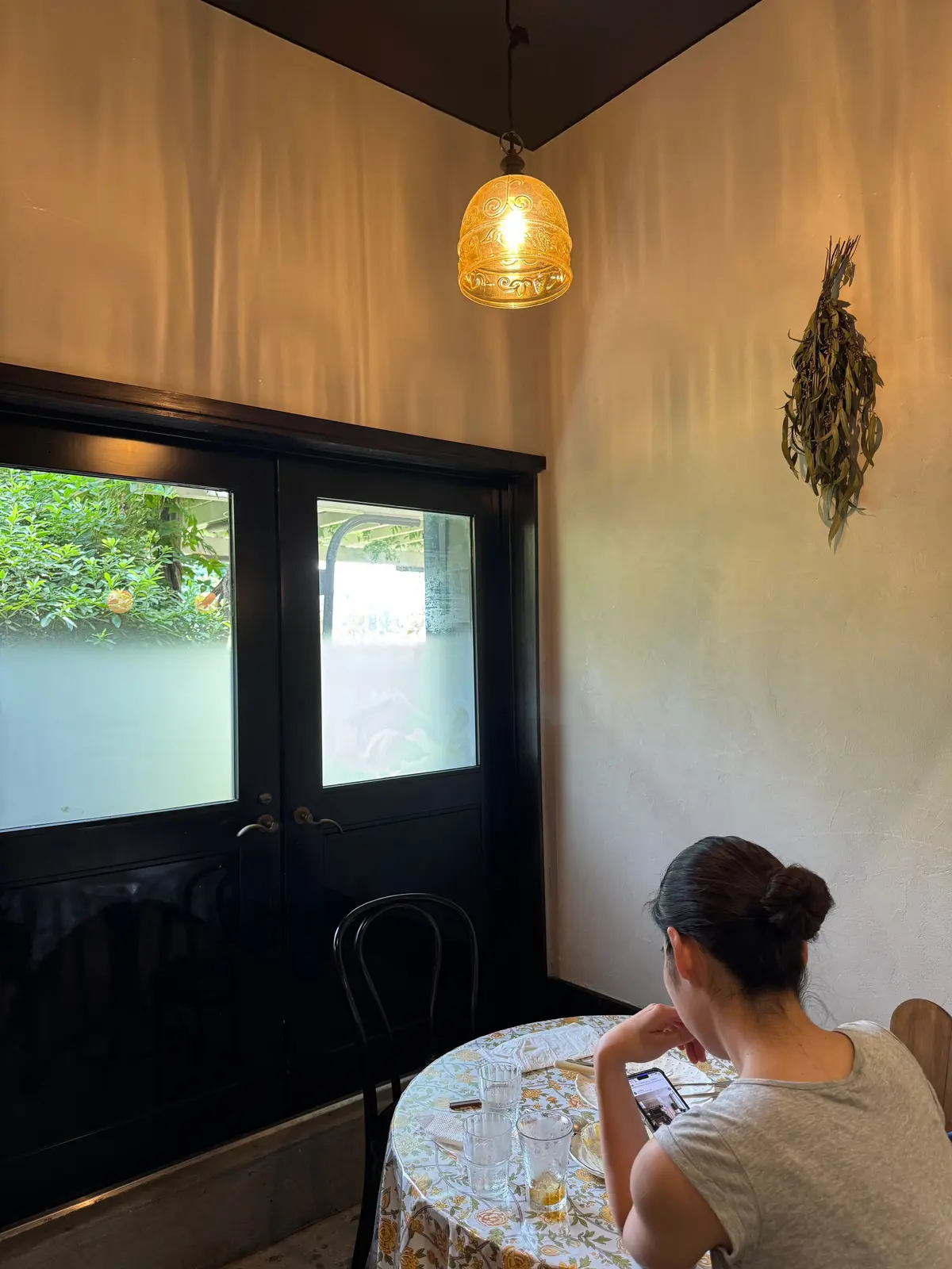 温かみのある部屋で女性がテーブルに座り、スマートフォンを見ている。テーブルには複数のグラスと小物があり、壁にはハーブの装飾とペンダントライトが設置されている。窓の外には緑の植物が見える。