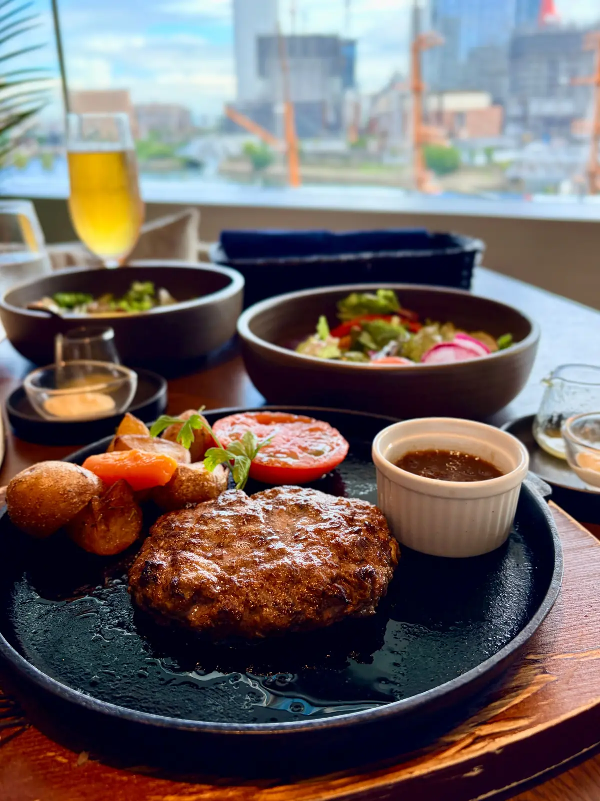 木製のテーブルに置かれた、ハンバーグ、サラダ、ビール、付け合わせの料理。ハンバーグは黒い皿に盛り付けられ、隣にはトマトとソース。背景には高層ビル群が見える。