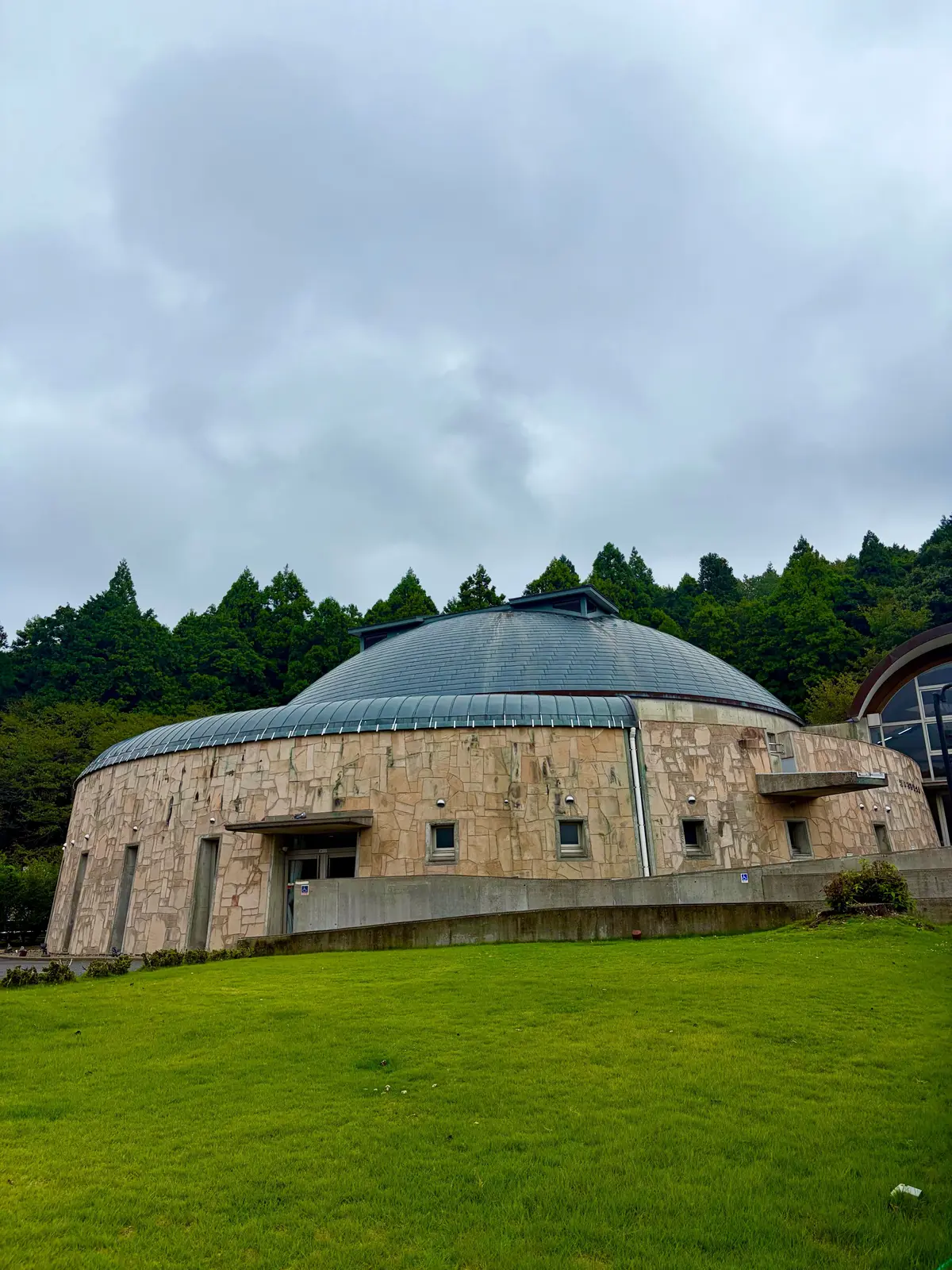円形に石材で覆われた建物。屋根は暗いグレーで、周囲に緑の芝生と木々が茂る。曇り空の下、建物は自然に溶け込んでいる。