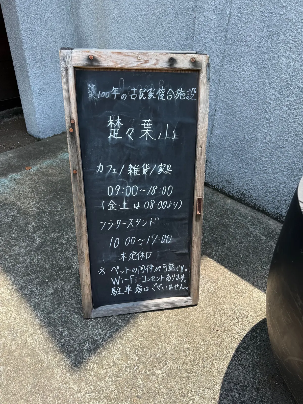 石造りの壁に立てかけられた木製の黒板に、手書きの文字で店の情報が書かれている。店名は「楚々菓山」、カフェ・雑貨・家具の営業時間は9時から18時（金土は8時から）、フラワースタンドは10時から17時。Wi-Fiあり、駐車場なしと記載されている。