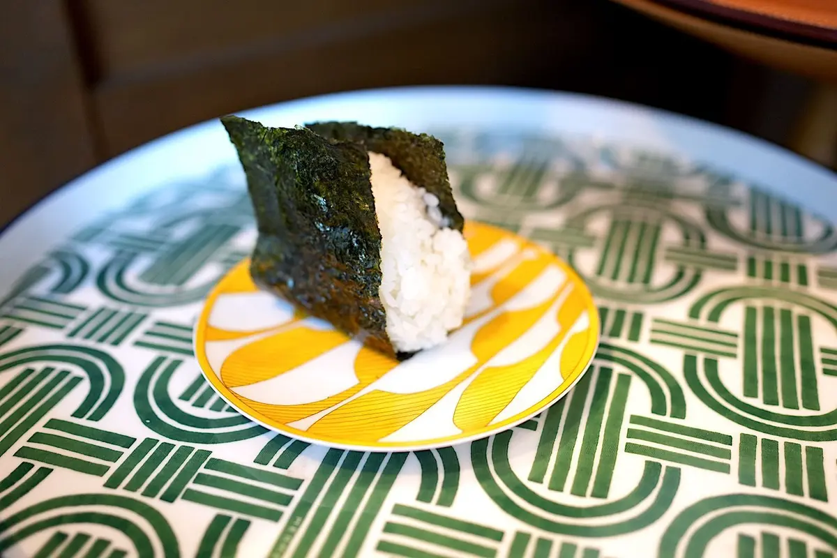 白い皿に置かれた、海苔で巻かれたご飯と、オレンジ色のソースをかけたお寿司。海苔は濃い緑色で、ご飯は白。皿には緑と白の幾何学模様が描かれている。