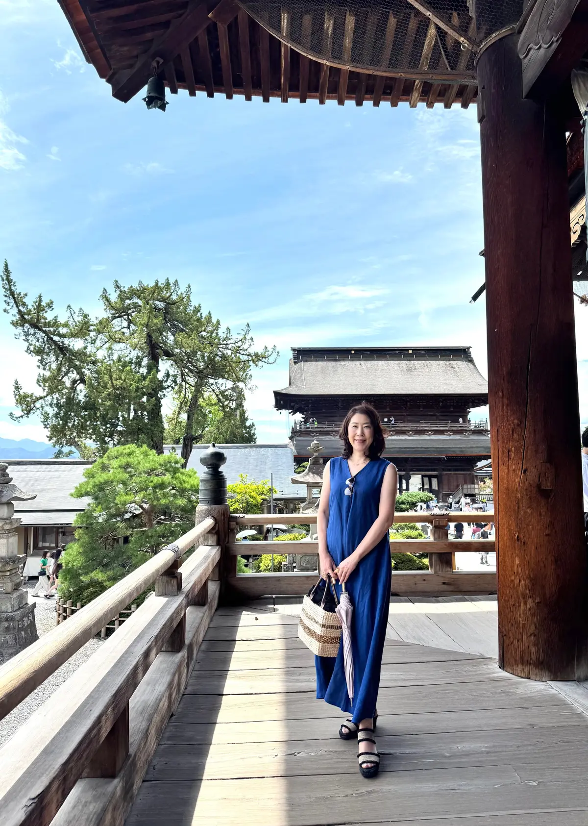 木造の回廊に立つ女性。青いワンピースを着て、麦わらのバッグを持ち、穏やかな表情。背景には、伝統的な日本の寺院建築と青空が広がる。