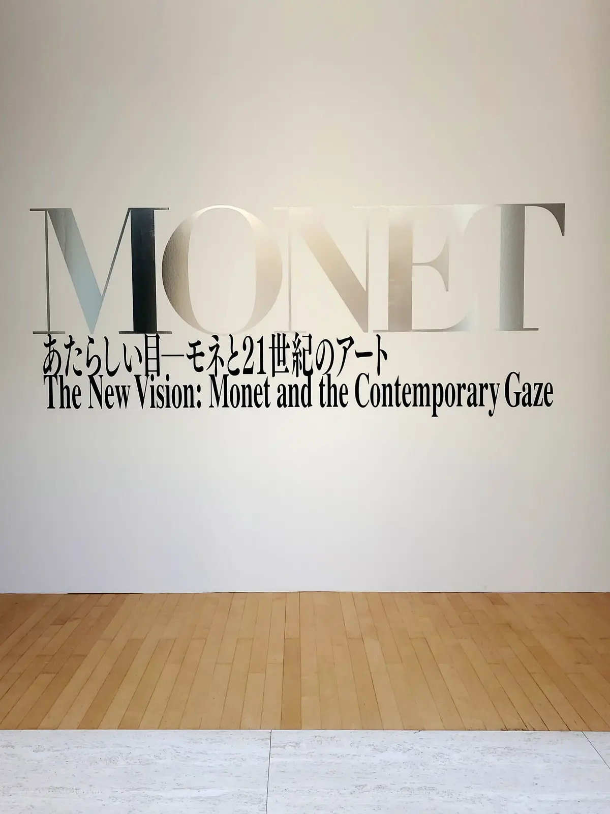 白い波板でできた立方体の建物に、「POLA MUSEUM OF ART」の文字が縦横に配置されている。建物の下には白と紫のストライプのパラソルが置かれている。背景には緑豊かな木々が見える。