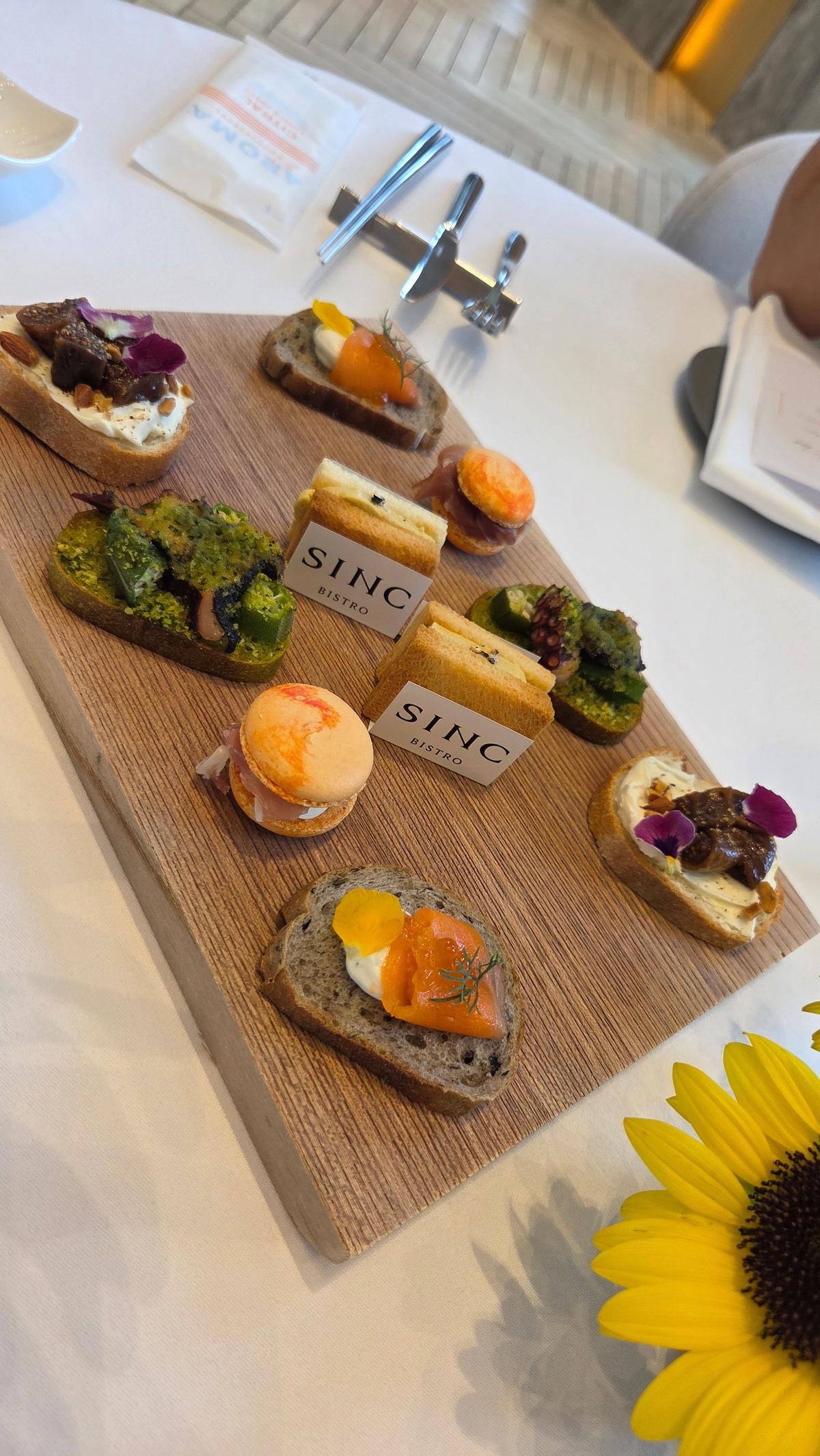 木製プレートに、SINC Bistroのロゴが入ったカードを挟んだ、様々なミニサンドイッチが並んでいる。エビ、クリームチーズ、卵、野菜を使ったものが、彩り豊かに配置されている。テーブルにはシルバーの食器と、白い紙、ひまわりの花が置かれている。
