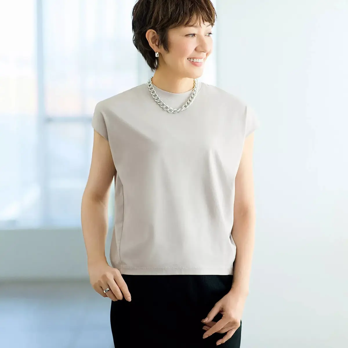 グレーのカットソーを着用した女性が微笑んでいる。肩幅が広く、短い袖口が特徴的。シルバーのネックレスと指輪を身につけ、黒いパンツと白い背景に立っている。