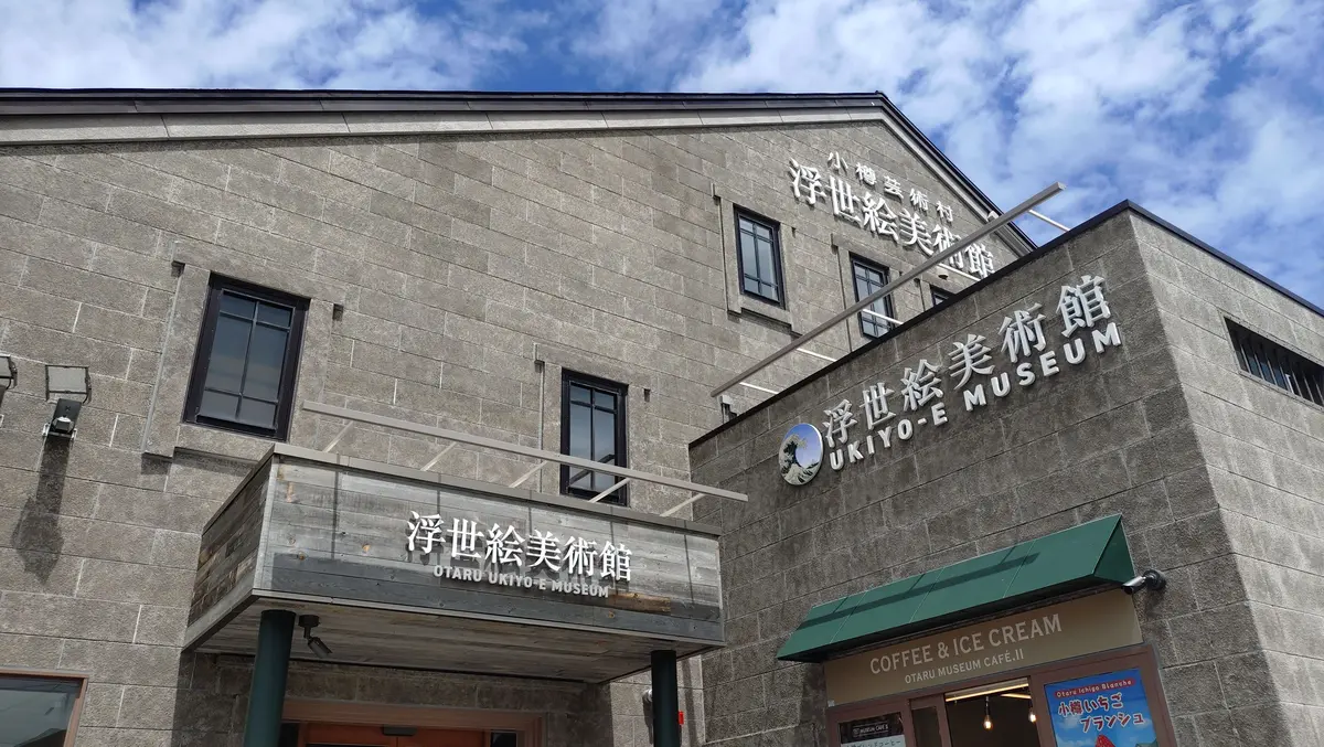 木造の灰色がかった建物で、屋根には「浮世絵美術館」の看板が掲げられている。入り口にはカフェの案内板があり、窓からは展示が見える。背景には山々が連なっている。