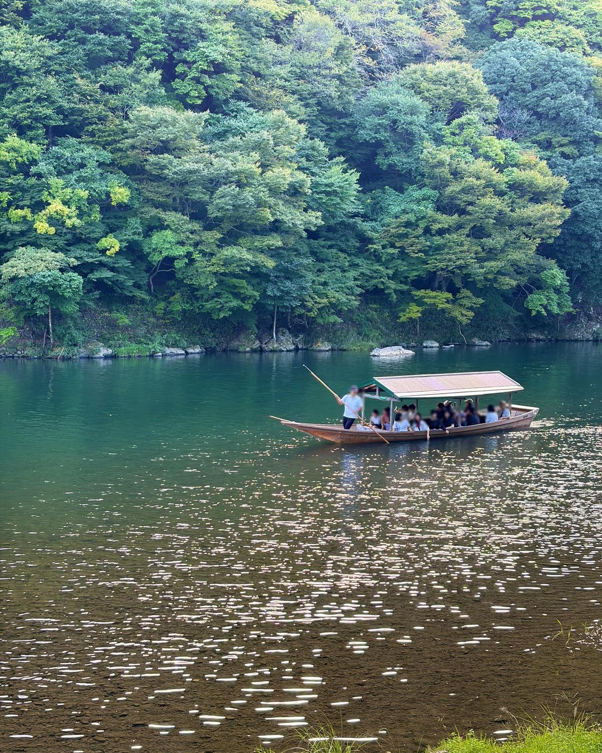 緑豊かな森林に囲まれた湖面を、乗客を乗せた伝統的な木製のボートが航行している様子。水面には光が反射し、キラキラと輝いている。