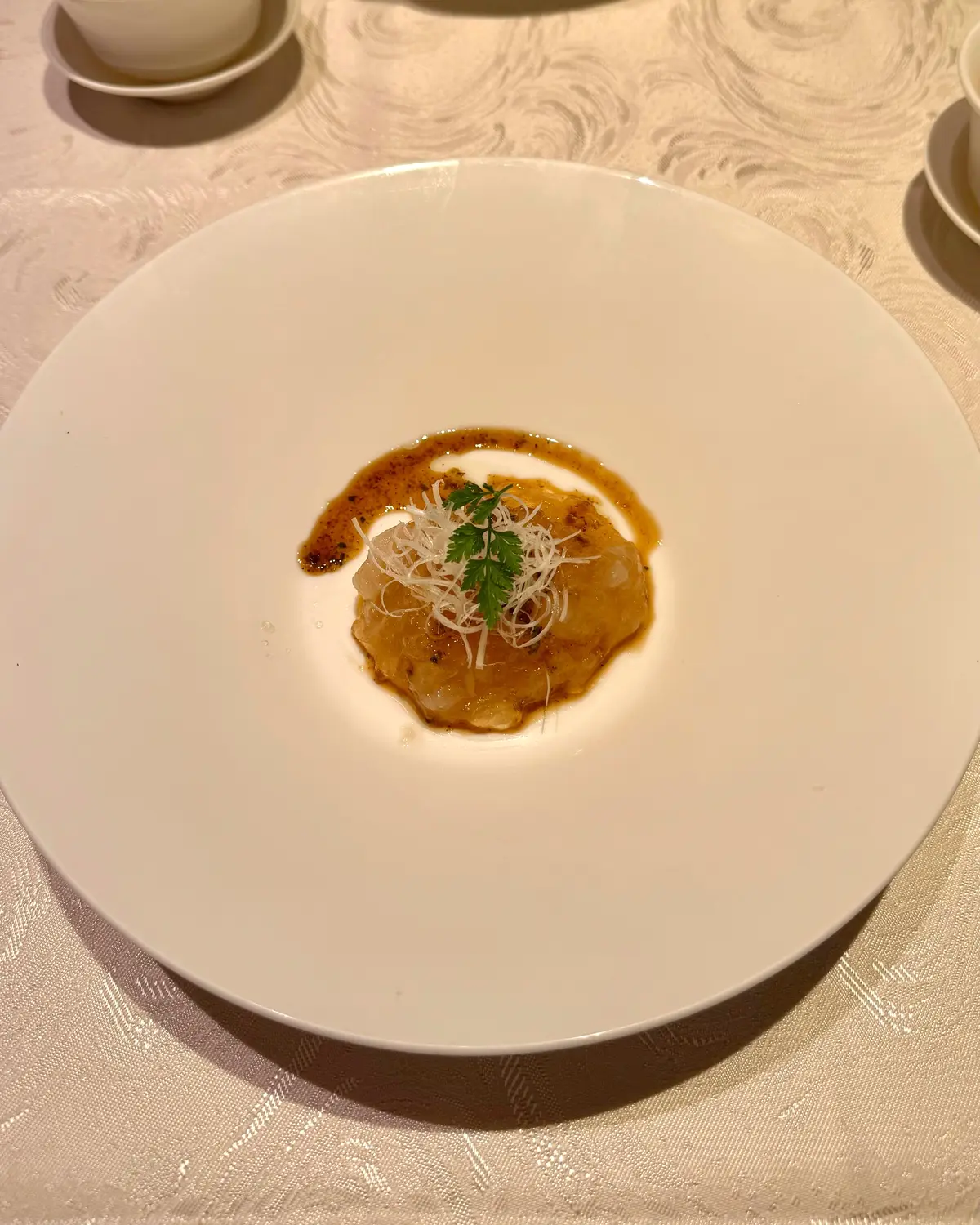 白い楕円形プレートに、黄色のクリーム状の料理が中央に盛り付けられ、周囲に茶色いソースが広がり、薄く削ったチーズと緑のハーブがトッピングされている。