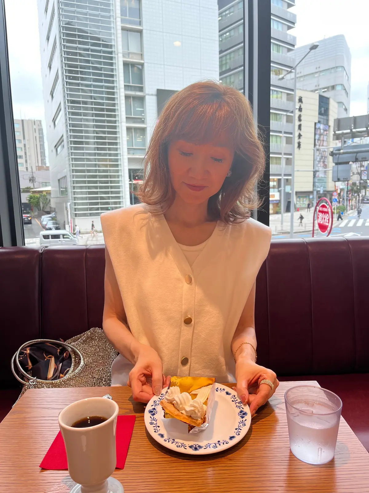 レストランの窓際の席で、女性がケーキを手に、少し困ったような表情をしている。茶色のショートヘアで、白いトップスを着用。窓の外には、高層ビル群と道路が見える。