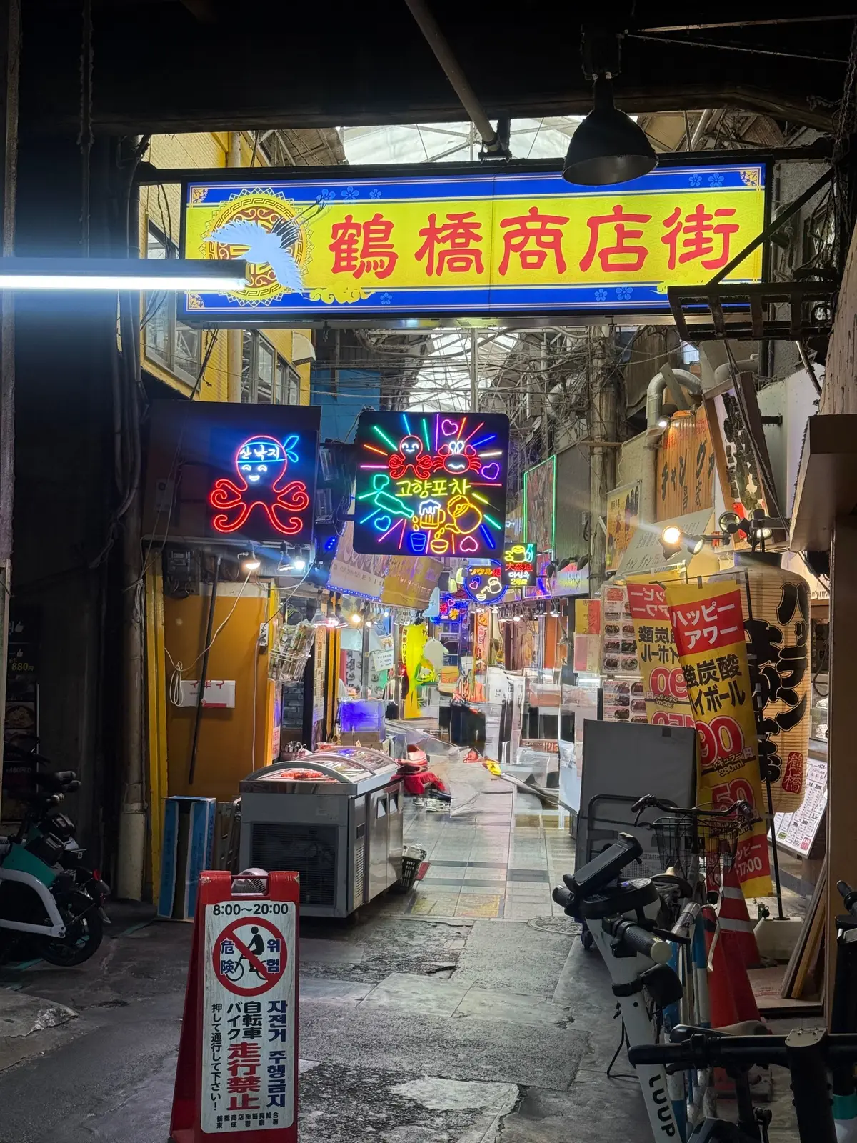 鮮やかなネオン看板が並ぶ活気ある商店街で、青と赤の文字が目立つ「亀橋商店街」の看板が中央に設置されている。左右にカラフルなネオンサインがあり、店頭には商品や自転車が置かれている。