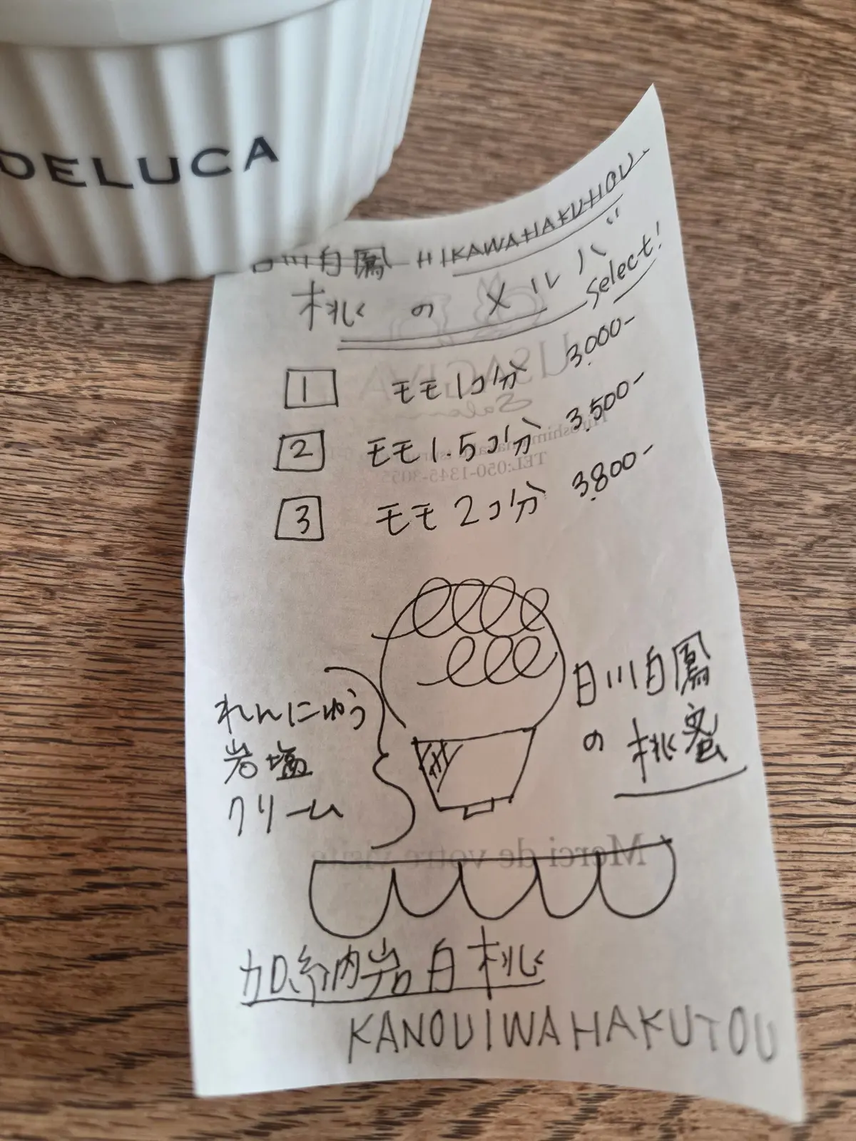 白いDELUCAのカップと、木製のテーブルに置かれた手書きのメモ。メモには、料金とイラストが描かれており、「日の出月闇の柿食」という文字が見える。