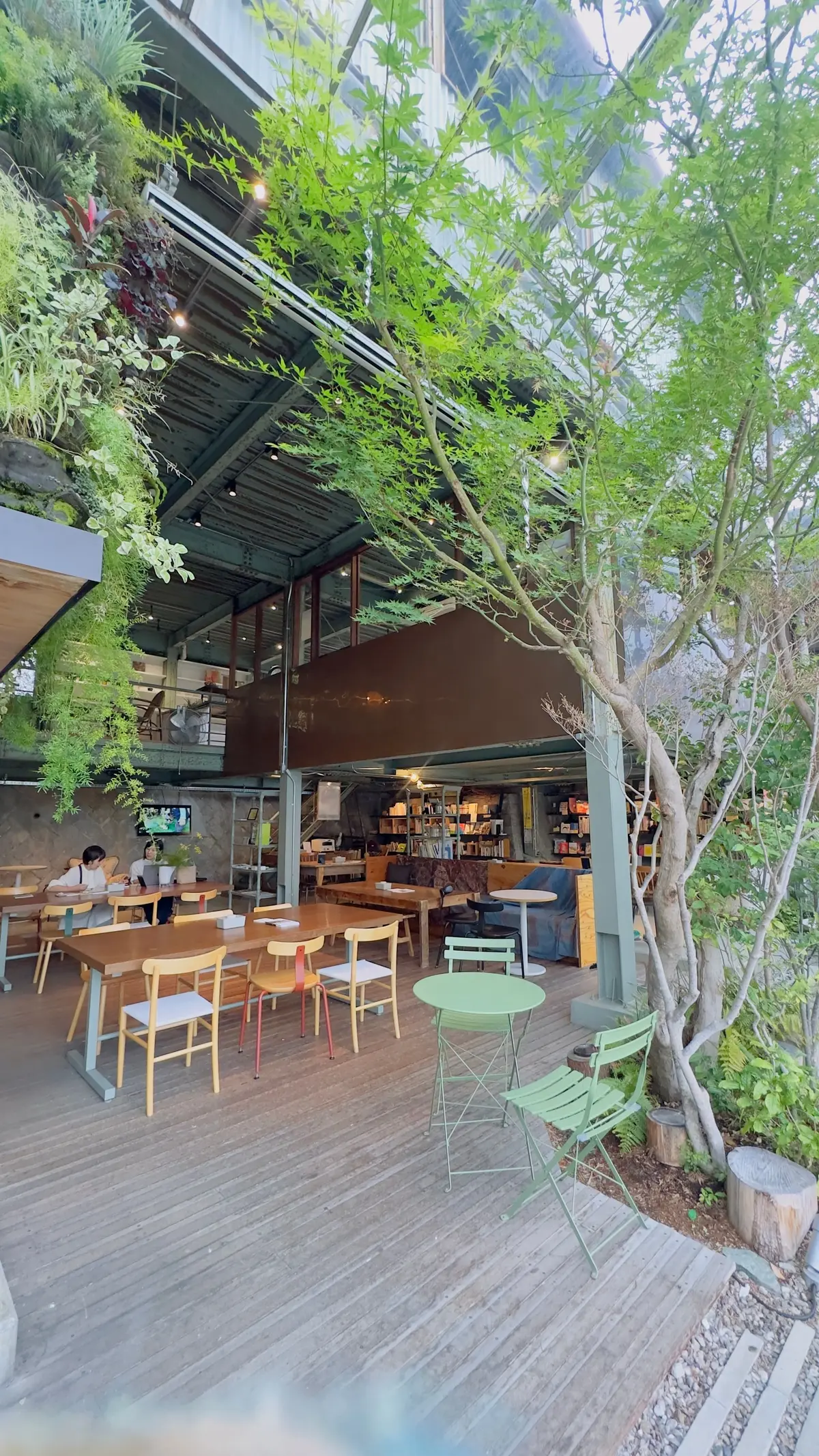 木製のテラスに配置されたテーブルと椅子、緑豊かな植物と本棚が並ぶ屋外カフェ