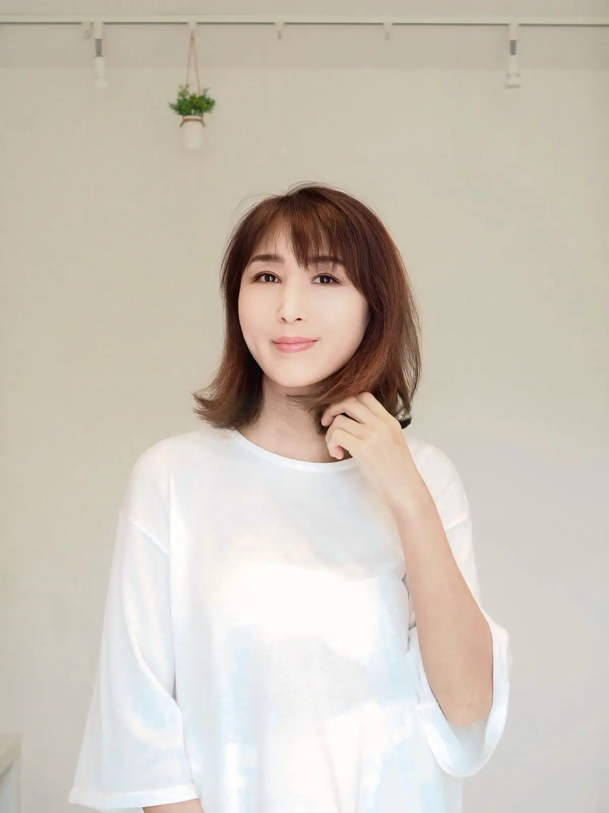 白いTシャツを着た女性が、明るい背景の前で微笑んでいる。茶色のボブヘアで、片手で髪を軽く触っている。