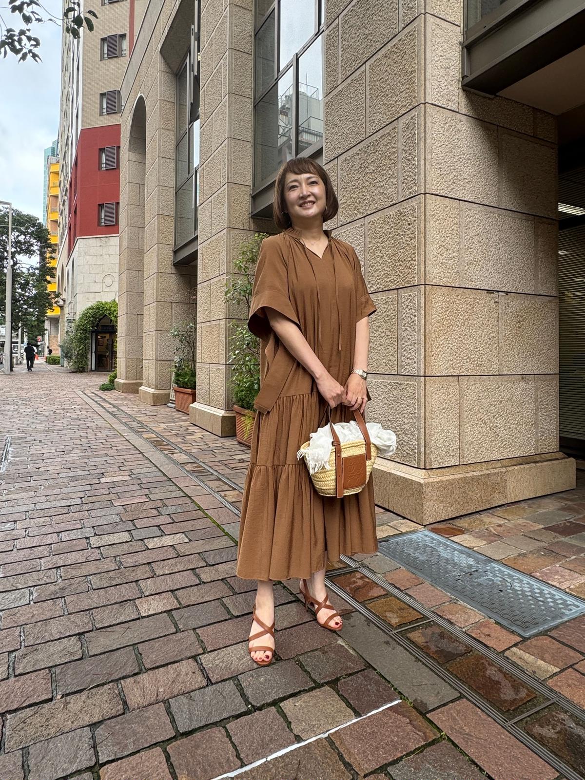 ブラウンのトップスとワンピースのセットアップを着た女性が、街路で微笑みながら立っている。茶色のサンダルを履き、同系色のカゴバッグを持つ。背景には、ベージュの建物と赤いアーチ型の窓が見える。